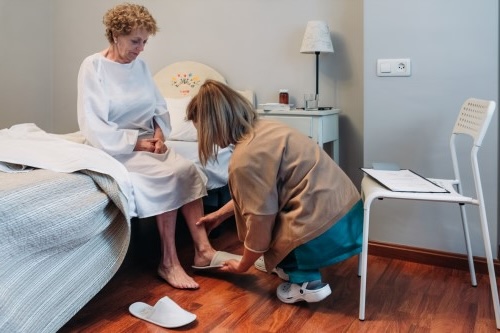 Carer helping elderly lady put on slippers on bed